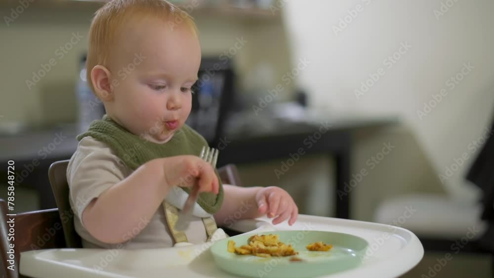 the one year old boy is sitting in the dining room and learning to eat with a fork himself the one year old boy is sitting in the dining room and learning to eat with a fork himself