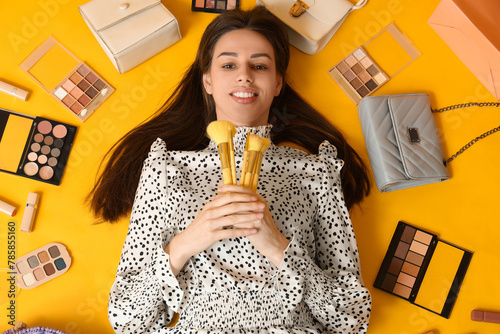 Happy smiling young woman with makeup brushes  eyeshadows palettes and handbags lying on yellow background