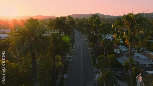 Aerial Establishing Shot of Sunlit Beverly Hills Neighborhood at Sunset, Golden Sun Above and Upscale Homes Below, Cars Driving Along Iconic Street with Mountains on Horizon