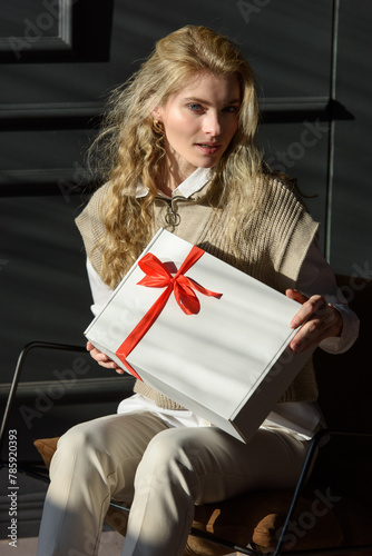 A woman in a chair holds a white gift box with a red ribbon