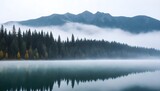 Trees partially obscured by fog on the edge of a calm lake, surrounded by mountains in the distance