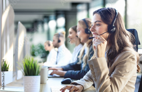 Female customer support operator with headset and smiling, with collegues at background.