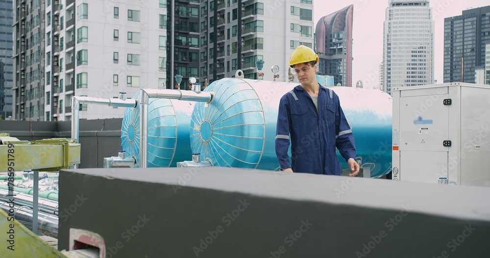 Engineer man in a blue jacket is walking inspecting maintenance insulated pipelines valve pump control on the roof at an industrial site near a large blue tank. Engineer man in a blue jacket is walking inspecting maintenance insulated pipelines valve pump control on the roof at an industrial site near a large blue tank.