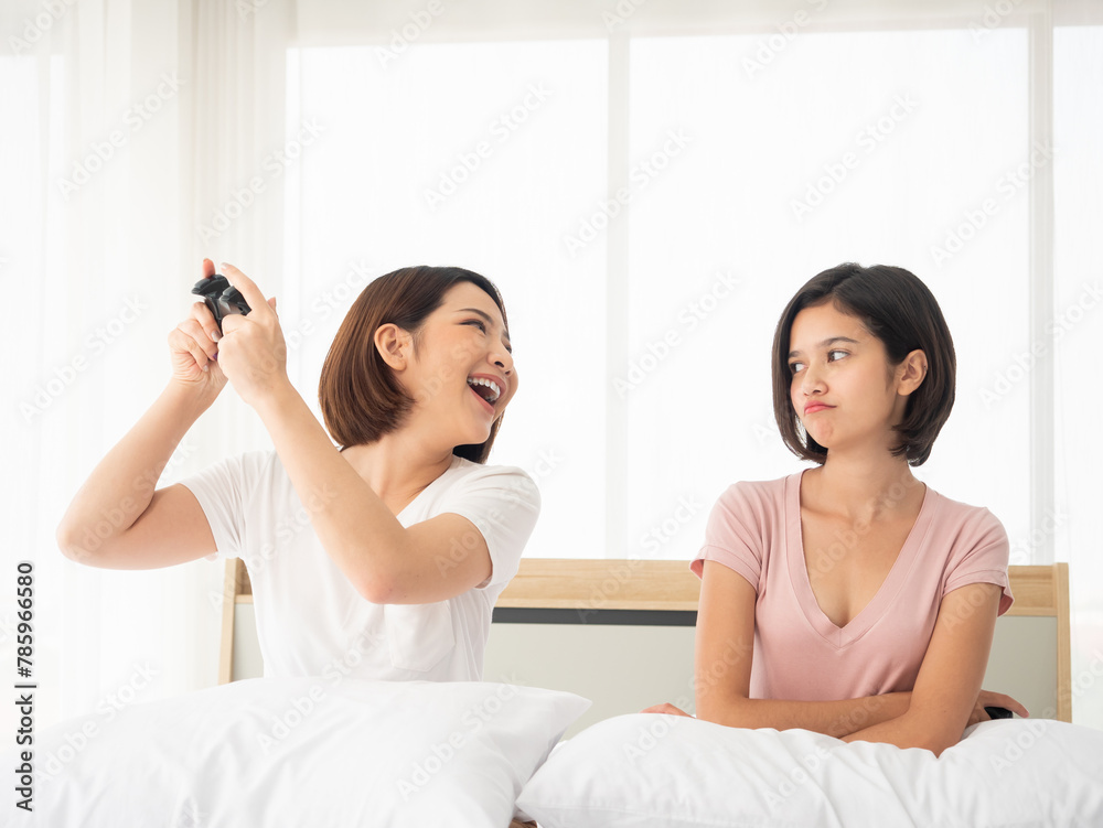 Asian two friends having fun in a bedroom, One gamer girl excitedly holding a game controller and laugh while the other angry with grumpy face Asian two friends having fun in a bedroom, One gamer girl excitedly holding a game controller and laugh while the other angry with grumpy face