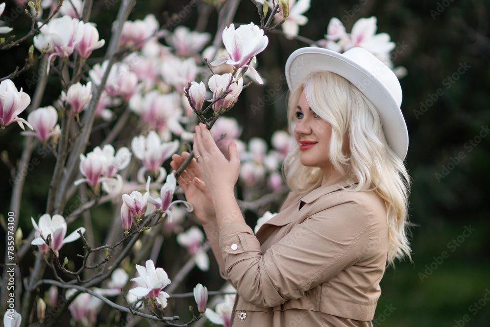 Naklejka premium Magnolia flowers woman. A blonde woman wearing a white hat stands in front of a tree with pink flowers. She has a smile on her face and she is enjoying the beautiful scenery.