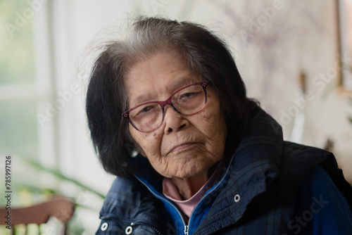 An elderly woman looks seriously at the camera. She is leaning to the side, with a warm vest on. Her hair has white roots, showing her grayness. She is Ryukyuan, from Okinawa, and in her nineties. photo