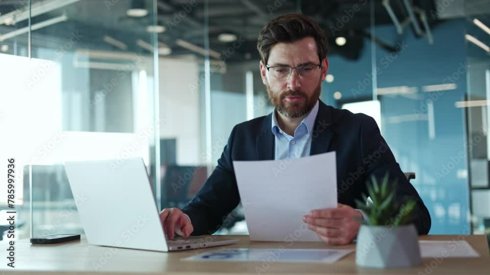Front view of cheerful businessman rewriting information from piece of important paper in wireless laptop in personal cabinet. Handsome bearded male engaging in paperwork with modern gadgets indoors. Front view of cheerful businessman rewriting information from piece of important paper in wireless laptop in personal cabinet. Handsome bearded male engaging in paperwork with modern gadgets indoors.