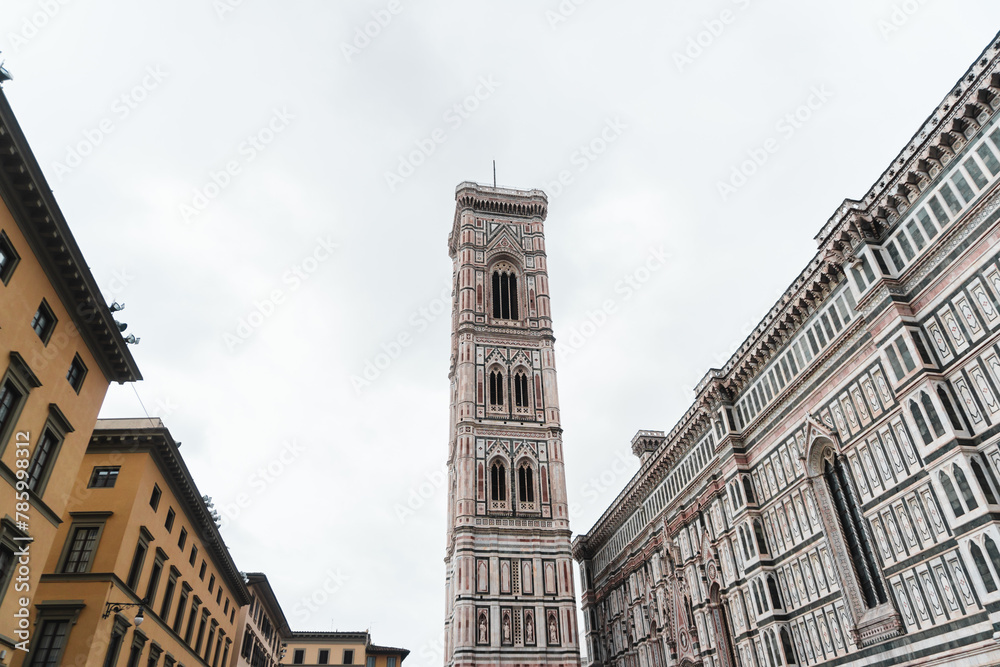 Medieval buildings and Santa Maria del Fiore's Gothic-Renaissance intricate façade framing Giotto's Campanile from Piazza del Duomo, standing tall in the centre, against the cloudy sky.