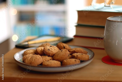 Cup of tea or coffee, pile of books, plate of cookies, reading glasses, e-reader and pen on the table. Colorful rainbow bookshelf in the background. Selective focus.