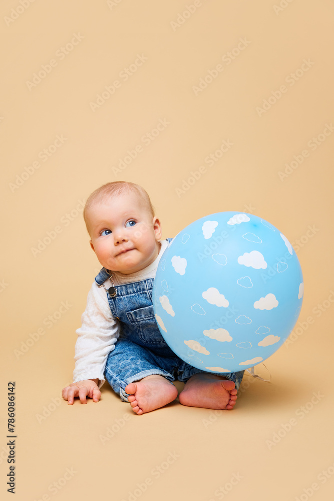 Cheerful infant, cute baby-girl in classic overalls laying with blue balloon against beige color studio background. Concept of childhood, motherhood, life, birth. Copy space for ad Cheerful infant, cute baby-girl in classic overalls laying with blue balloon against beige color studio background. Concept of childhood, motherhood, life, birth. Copy space for ad