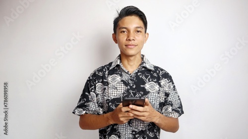 Happy young handsome Asian man wearing batik shirt posing holding smartphone photo