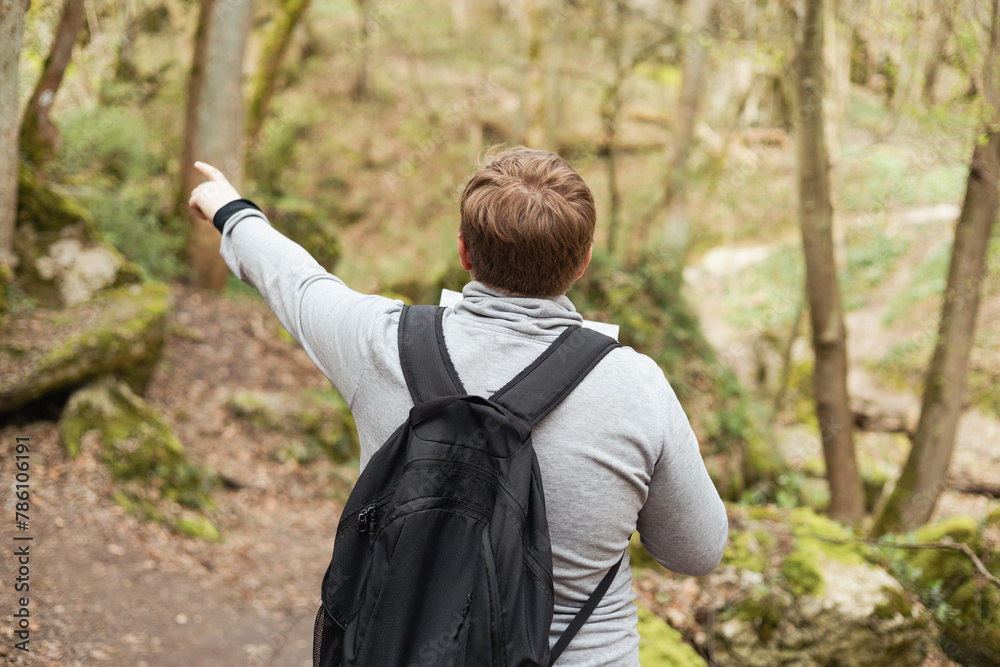 Young men with paper map walking in forest. Blonde male in sport clothes with backpack pointing finger outdoors, he hiking on woodland, rear view Young men with paper map walking in forest. Blonde male in sport clothes with backpack pointing finger outdoors, he hiking on woodland, rear view