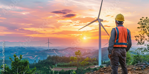 Engineer wearing an hardhat stands on top of a hill and looks at a beautiful sunset windmill landscape