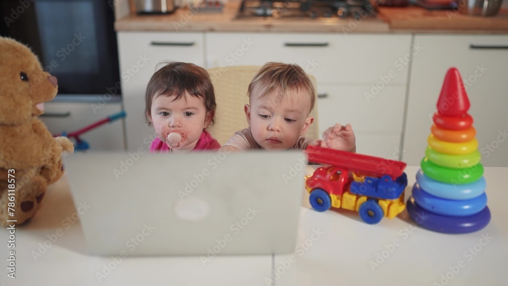 Fototapeta premium twin baby children a playing laptop watching video in the kitchen. happy family kid dream concept. baby twins lifestyle playing video game on laptop looking at screen in kitchen