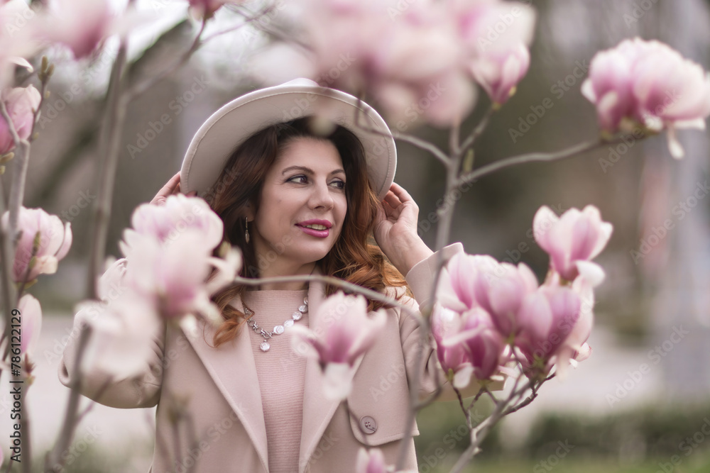 Fototapeta premium Woman magnolia flowers, surrounded by blossoming trees., hair down, white hat, wearing a light coat. Captured during spring, showcasing natural beauty and seasonal change.