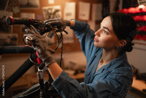 Close-up portrait of concentrated cycling mechanic woman checking and repairing bicycle handlebar with tools while working in bike repair workshop with authentic interior.