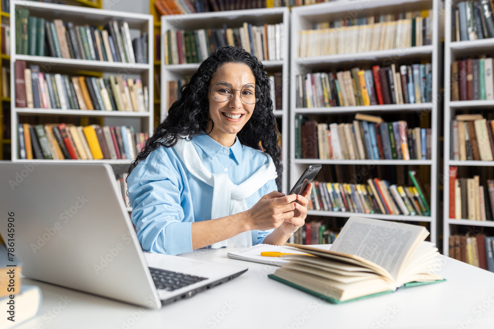 A cheerful woman wearing glasses interacts with her smartphone while seated in a library surrounded by books. She exudes a sense of happiness and content in an academic setting. A cheerful woman wearing glasses interacts with her smartphone while seated in a library surrounded by books. She exudes a sense of happiness and content in an academic setting.
