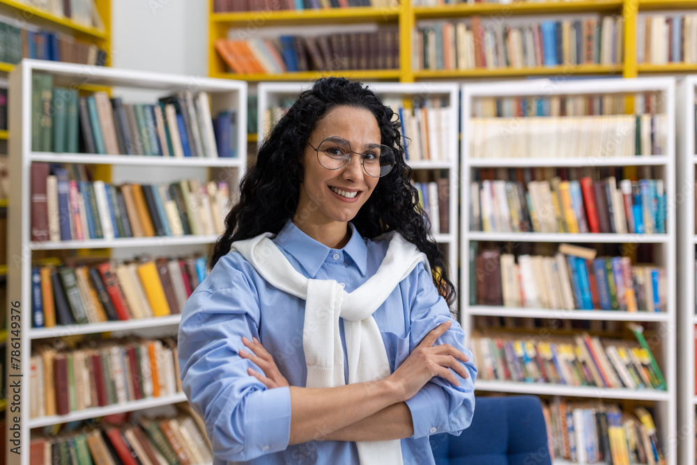 Portrait of a confident, smiling female librarian standing with arms crossed in a brightly colored library full of books. She wears glasses and a casual outfit.