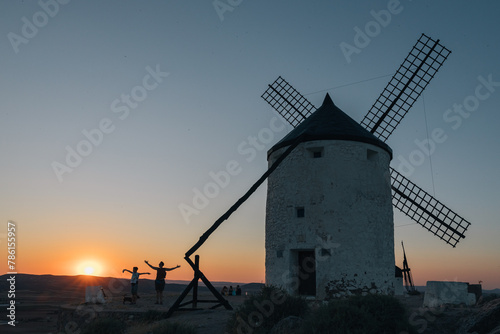 A windmill is standing in the distance with the sun setting behind it, two unrecognizable persons open their arms, the scene is peaceful and serene, with the sun casting a warm glow on the landscape