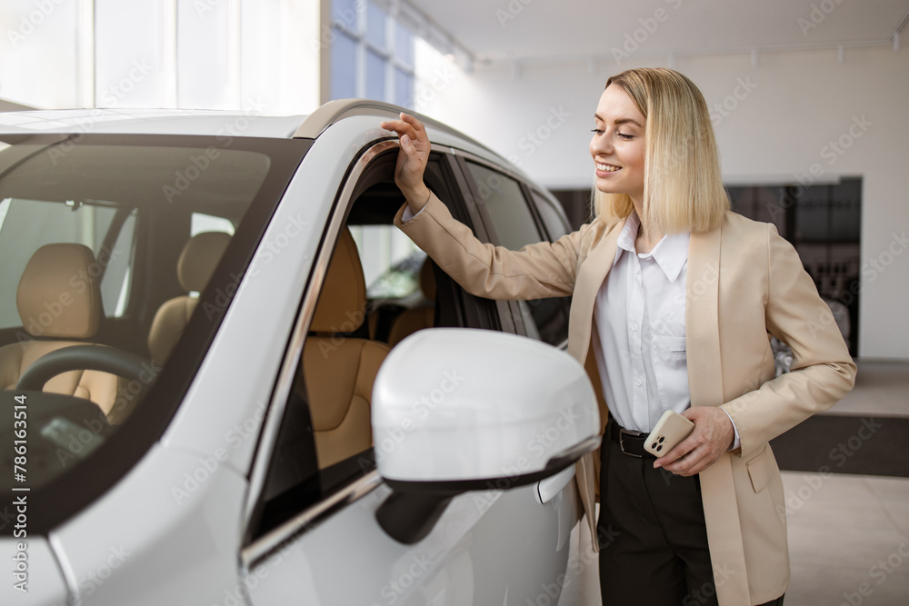 Happy, fun woman customer buyer, client, wearing yellow suit, looking at interior of car through lowered window. Blond young female choosing auto, wants to buy new automobile in car showroom.