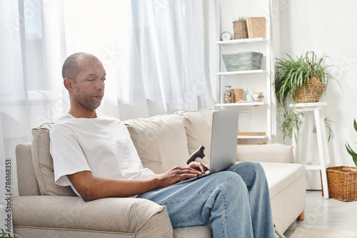 An African American man with myasthenia gravis sits on a couch, using a laptop computer