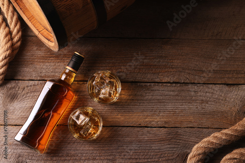 Whiskey with ice cubes in glasses, bottle and barrel on wooden table, flat lay. Space for text