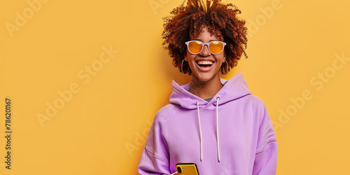 Happy African millennial woman laughing with mobile phone on yellow studio backdrop. Joyful black girl wearing purple hoodie, sunglasses smiling looking camera. Great new mobile App. People and gadget
