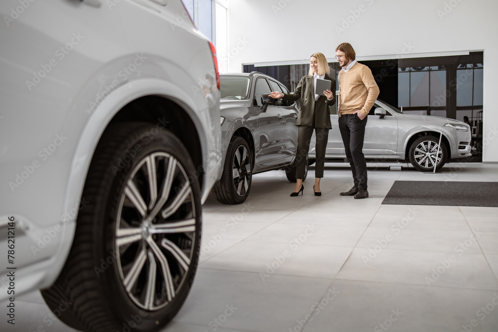 Caucasian male customer in vehicle showroom, female person selling transport, auto dealer business. Confident blond saleswoman explaining all the car features using tablet to handsome owner.
