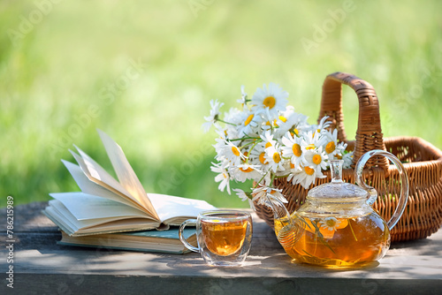 glass teapot and cup with herbal tea, Chamomile flowers in basket, books on table in garden. Summer nature background. relax, reading time. useful calming tea. tea party