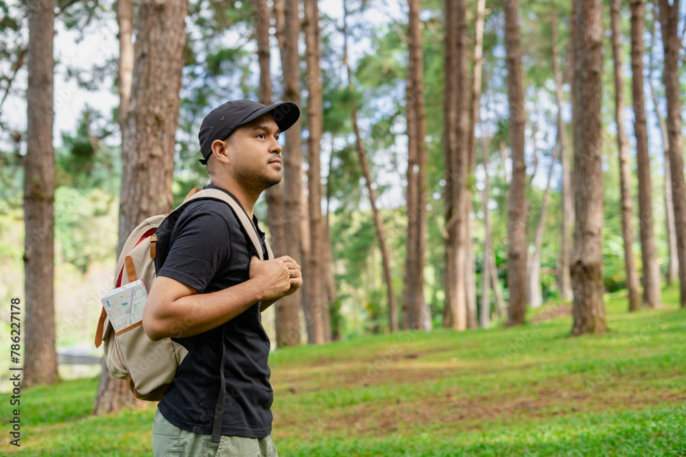 Fototapeta premium Young happy man standing and looking at distance enjoying the fresh air in the forest. Male hiker with backpack and map with open arms enjoying and relax the nature.
