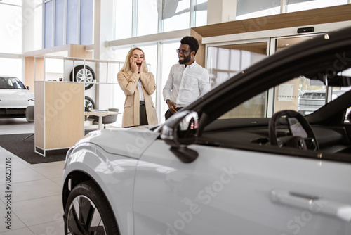 Happy husband buying to his loving wife new vehicle at car dealership. African American boyfriend covering eyes of his Caucasian young girlfriend for surprise in car showroom. © sofiko14