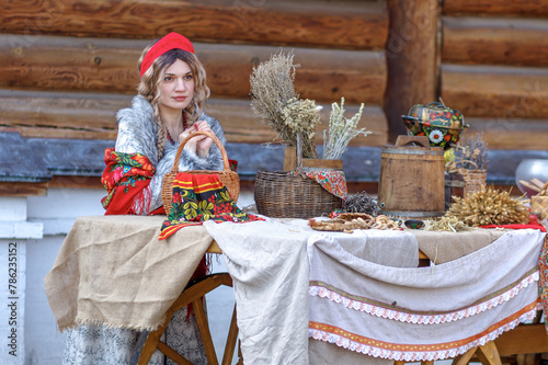 A beautiful Russian girl in traditional Russian clothes and a kokoshnik stands at a table with various national dishes near a wooden house. Festive farewell to winter. photo