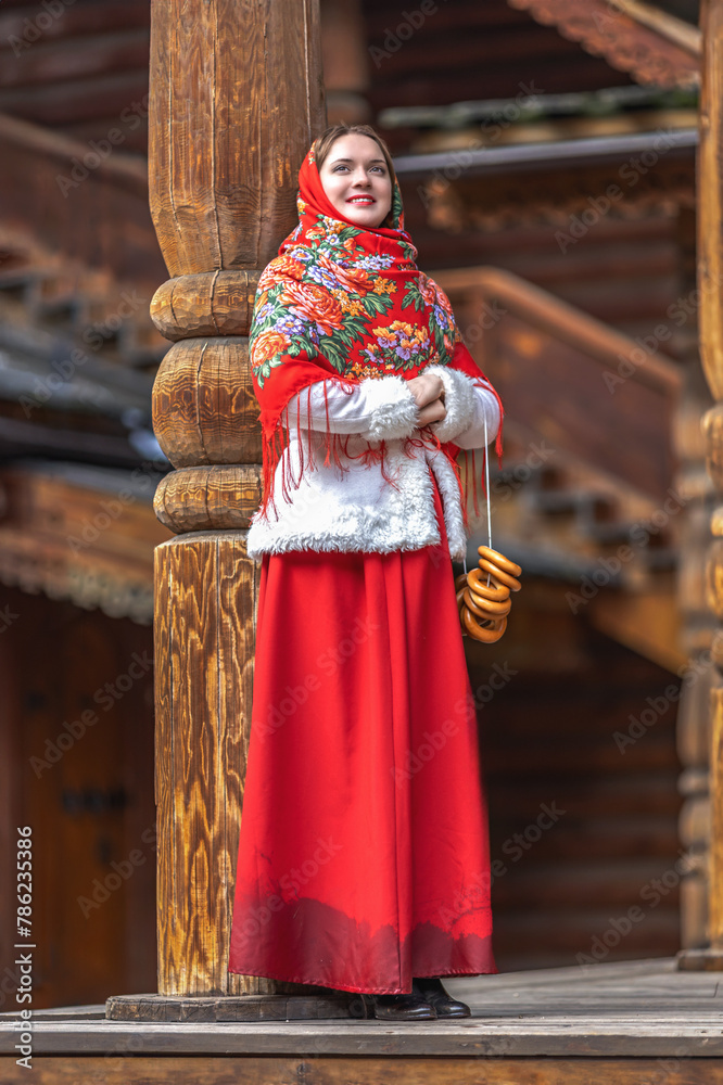 A beautiful Russian girl in red traditional Russian clothes holds bagels in her hands in the spring near a wooden house. Festive farewell to winter. A beautiful Russian girl in red traditional Russian clothes holds bagels in her hands in the spring near a wooden house. Festive farewell to winter.