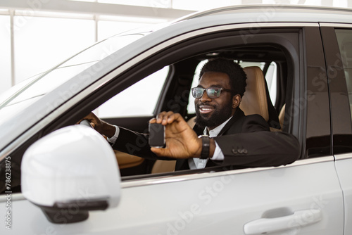 Auto business, car sale, consumerism and people concept. Happy African man taking car key from dealer in bright showroom or salon and showing them to camera. © sofiko14