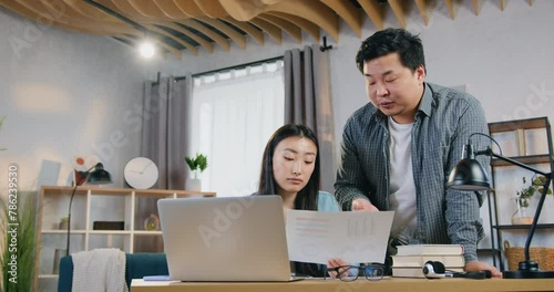 Attractive asian man standing near woman and helping with university hometask on computer using report with charts photo