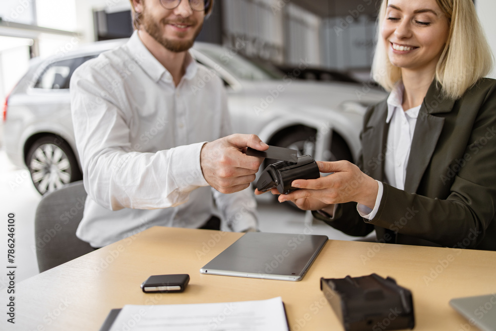 Obraz premium Female confident manager helps to male customer make payment for purchase of branded crossover. Happy young man paying with electronic credit card at terminal for purchase of new modern car.