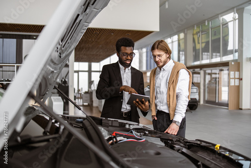 Profession consultant service showing choosing buying a car. Young Caucasian male customer and professional African car dealer standing near new car with open hood examining engine of vehicle.
