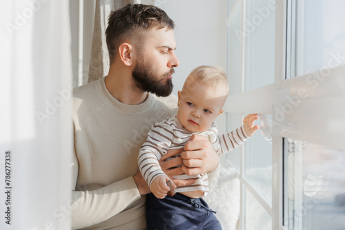 Heartwarming scenes of bearded father in beige sweater bonding with his cheerful baby son, enjoying sunny comfort of home setting. Concept lifestyle moment in life of fatherhood © Parilov