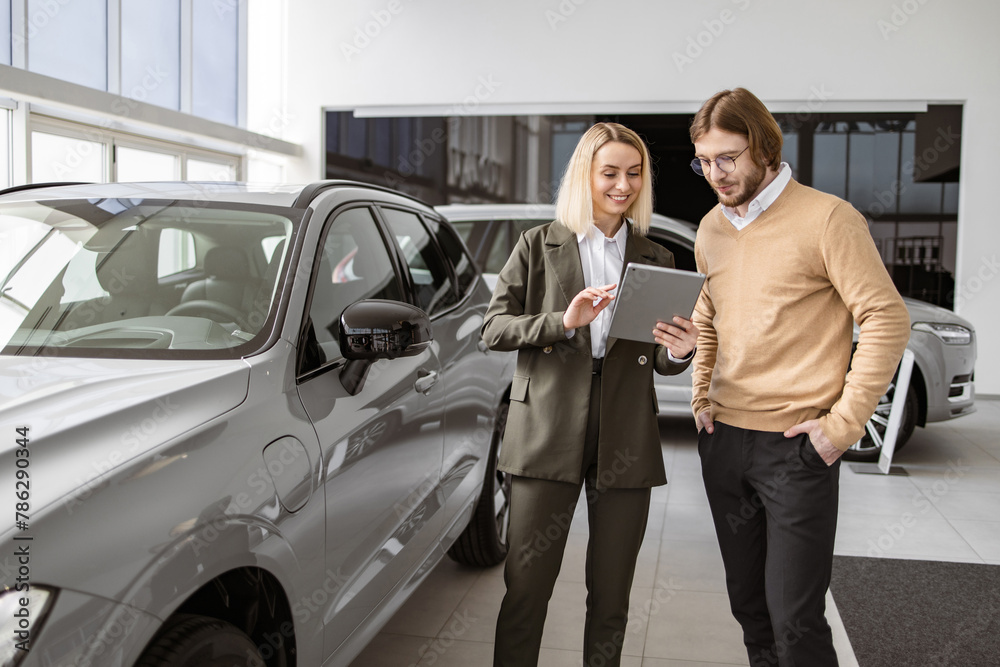 Caucasian male customer in vehicle showroom, female person selling transport, auto dealer business. Confident blond saleswoman explaining all the car features using tablet to handsome owner.