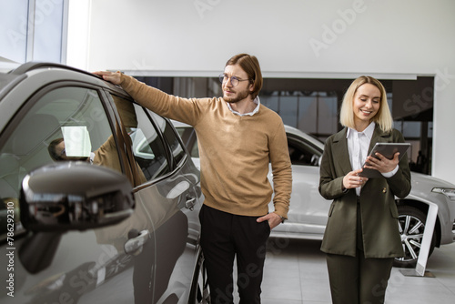 Successful businessman in car showroom, sale of vehicles to customers. Professional female consultant provides all information about new car using tablet to Caucasian male client at car dealership. © sofiko14