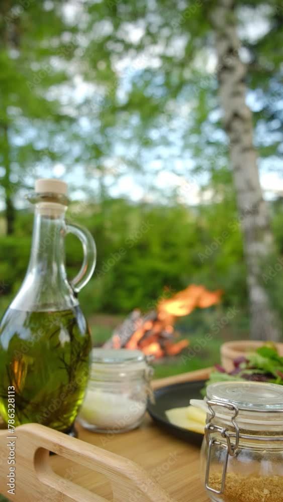 Vertical video of olive oil, salt, leaves salat, food to prepare on wooden board near bonfire. Plate of food in clearing on background campfire and forest at spring or summer day Vertical video of olive oil, salt, leaves salat, food to prepare on wooden board near bonfire. Plate of food in clearing on background campfire and forest at spring or summer day