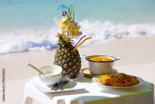 Asian beach still life with fresh exotic cocktail in pineapple decorated tropical flowers. Thai Tom Yum soup and Pad Thai noodles with cutlery on a small beach table along with a cocktail on the beach