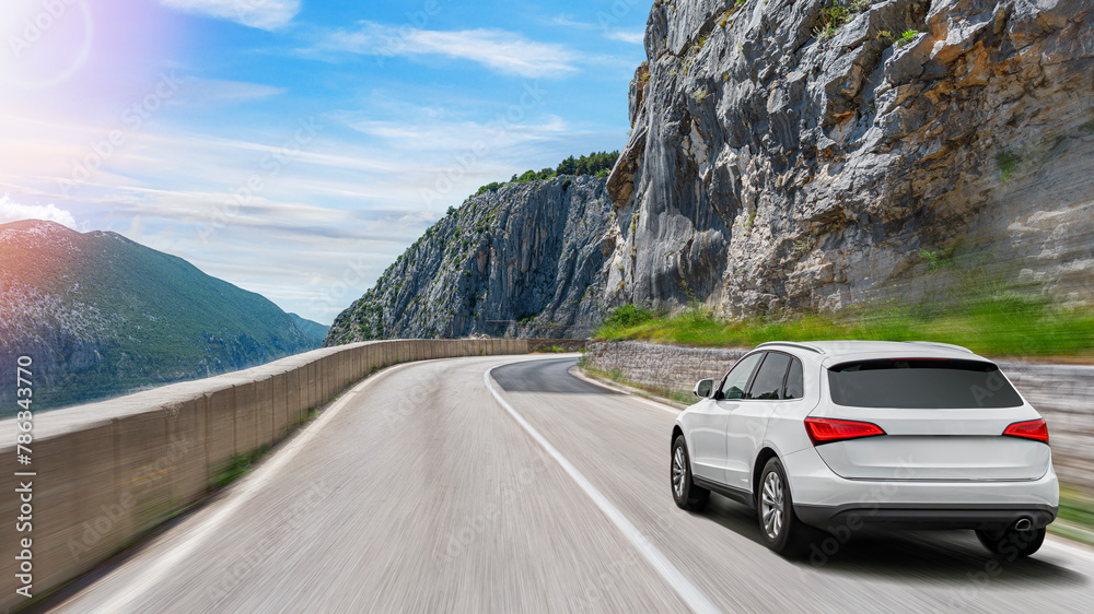 A white car drives along the highway against the backdrop of rocky mountains on a sunny day.