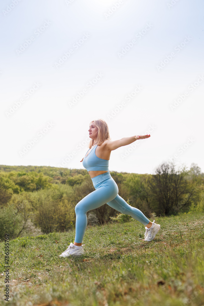The female athlete spreads her arms to the sides and performs lunges forward. Beautiful blonde Caucasian woman in blue tight tracksuit. Blonde girl at an outdoor training session The female athlete spreads her arms to the sides and performs lunges forward. Beautiful blonde Caucasian woman in blue tight tracksuit. Blonde girl at an outdoor training session