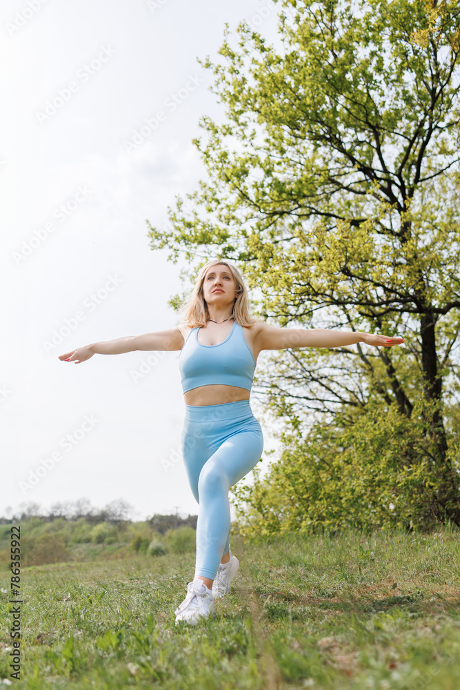 The female athlete spreads her arms to the sides and performs lunges forward. Beautiful blonde Caucasian woman in blue tight tracksuit. Blonde girl at an outdoor training session