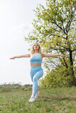 The female athlete spreads her arms to the sides and performs lunges forward. Beautiful blonde Caucasian woman in blue tight tracksuit. Blonde girl at an outdoor training session