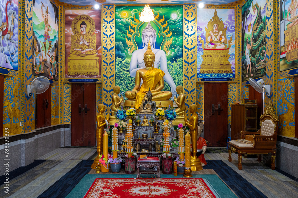 Chachoengsao, Thailand, 28 July 2023. Wat Khao Tham Raet, Golden Buddha statue sitting in a temple. The statue is seated on a red rug and has its eyes closed. Behind the statue is a mural on the wall.