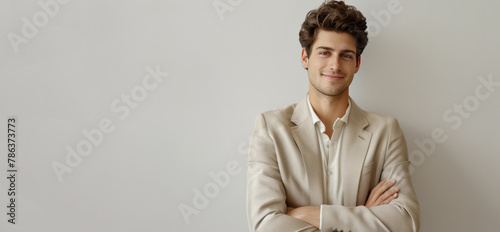 A young businessman in a light gold suit stands with his arms crossed, smiling against a silver backdrop. photo