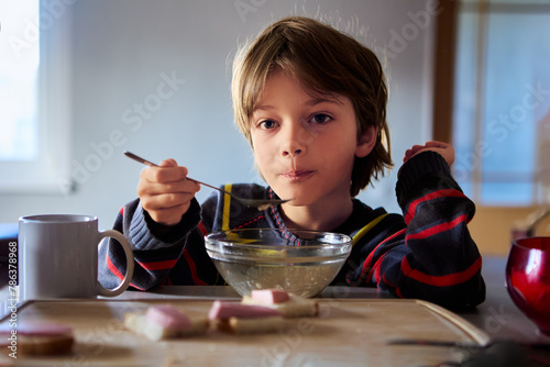 Unhappy male child sits in kitchen table with bowl of cereal in dimly lit kitchen. photo
