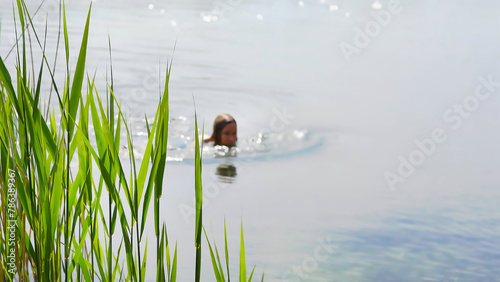 blurred swimming woman in a lake in summertime with focus on reed grass in foreground, idyllic natural background for travel, beach vacation and sports